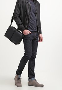 Svart bomberjacka över en textured marinblå skjorta, slim fit mörka jeans och grå ankelboots med ljusblå manschetter, hållande en svart crossbody-väska.