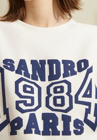 Valkoinen huppari, jossa on suuri laivaston sininen teksti "SANDRO 1984 PARIS." Kangas on tekstuuriltaan ja siinä on pyöreä kaula-aukko.