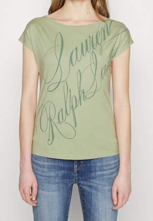 Kobieta ubrana w jasnozieloną koszulkę z krótkim rękawem z "Lauren Ralph Lauren" dużą kursywą i niebieskie dżinsy.