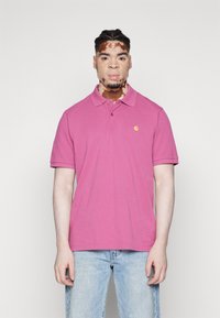 Camisa polo de algodón rosa con cuello y dos botones, que presenta un pequeño logo amarillo en el lado izquierdo del pecho, combinada con unos vaqueros celestes.