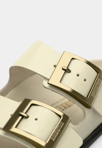 Ανοιχτό κίτρινα σανδάλια με δύο φαρδιές λουριά, χρυσές αγκράφες και πέλμα σε γυμνό χρώμα. Το υλικό φαίνεται λείο και επίπεδο.