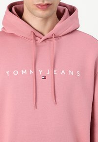Felpa con cappuccio rosa con cordini, con logo "TOMMYJEANS" bianco e un piccolo accento a forma di bandiera colorata sotto. Realizzata in morbido cotone.