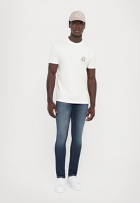 Fehér rövid ujjú póló bézs logóval a mellrészen, sötétkék, slim-fit farmernadrággal és fehér sportcipővel párosítva, bézs sapkával kiegészítve.