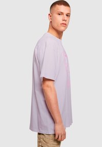 Světle fialové oversized tričko vyrobené z bavlny, s růžovým grafikou na přední straně, krátkými rukávy a kulatým výstřihem.