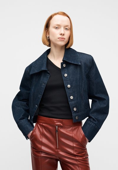 Νεαρό άτομο με κοντά κόκκινα μαλλιά που φοράει σκούρο τζιν μπουφάν, μαύρο τοπ και καφέ ψηλόμεσα δερμάτινα παντελόνια με φερμουάρ μπροστά.