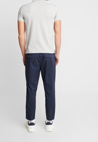 Homme debout, de dos, portant un polo à manches courtes gris clair, un pantalon bleu marine, des chaussettes blanches et des baskets blanches avec des accents bleus marine.