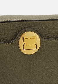 Borsa a tracolla in pelle testurizzata verde oliva con una chiusura circolare dorata. Presenta una struttura liscia e una cucitura delicata lungo il bordo.