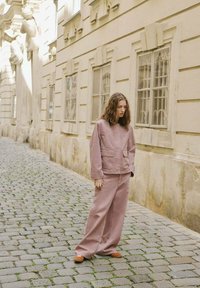 Giacca di cotone rosa con tasche frontali abbinata a pantaloni larghi coordinati. La modella si trova su una strada acciottolata vicino a un edificio pallido.