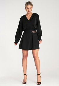 Czarna sukienka z dekoltem w serek, długimi rękawami i dopasowaną talią z brązowym paskiem. Spódnica jest plisowana i delikatnie się rozszerza, charakteryzując się gładką teksturą.