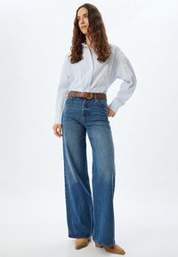 Camisa a rayas azul claro con mangas anchas, combinada con jeans de pierna recta de talle alto en azul oscuro, acentuada con un cinturón marrón y zapatos beige.