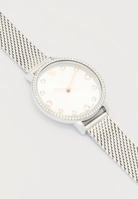 Ασημένιο ρολόι με λουράκι από μέταλλο πλέγμα, στεφάνη με μικρά κρύσταλλα, λευκό καντράν, ενδείξεις ωρών από κρύσταλλο και δείκτες ώρας και λεπτών σε χρυσό χρώμα.