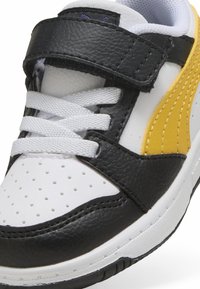 Fekete-fehér sneaker sárga kiegészítőkkel, texturált bőrből készült felsőrész, perforált orr résszel, állítható tépőzáras pánttal és fűzőkkel.