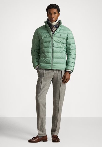 Nafukovaná zelená bunda so zipsom, nosená cez biely rolák. Kombinovaná so svetlosivými nohavicami a hnedými topánkami, doplnená o károvaný šál.