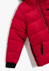 Rød quilted jakkeærme med sort manchet, lodret lynlås lomme med sort trim, og en matchende rød lynlås på en hvid baggrund.