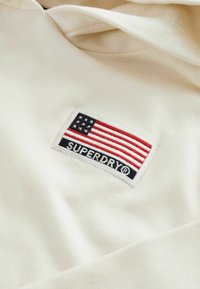 Krämfärgad hoodie med en rektangulär lapp som har en vertikalt randig amerikansk flagga och varumärkesnamnet "SUPERDRY" under.