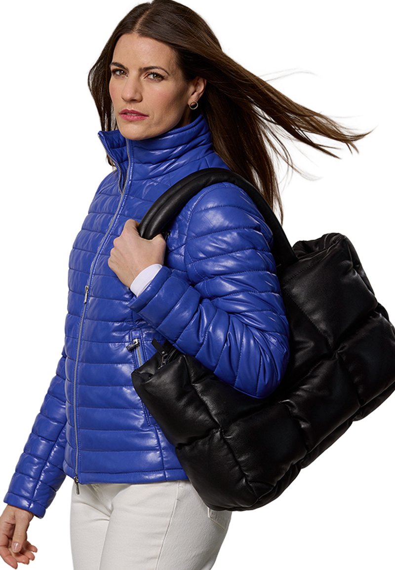 Blaue gesteppte Steppjacke, kombiniert mit einer schwarzen, gepolsterten Tragetasche. Das Modell hält die Tasche an einem eleganten schwarzen Griff.