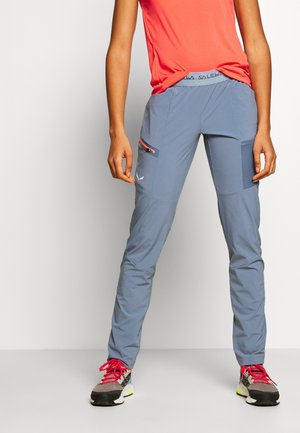 Persoon die een blauwe outdoorbroek draagt met een met ritssluiting afsluitbare dijbeenzak en grijze wandelschoenen met roze veters, staand tegen een effen achtergrond.