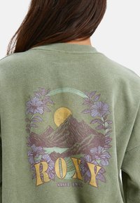Vrouw in een groene wafelgebreide trui met een berg, zon, paarse bloemen en "ROXY Sinds 1990" gedrukt op de achterkant.
