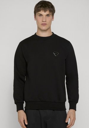 Ragazzo con capelli castani corti che indossa una felpa nera con collo a giro e un piccolo logo nero a forma di diamante sul petto.