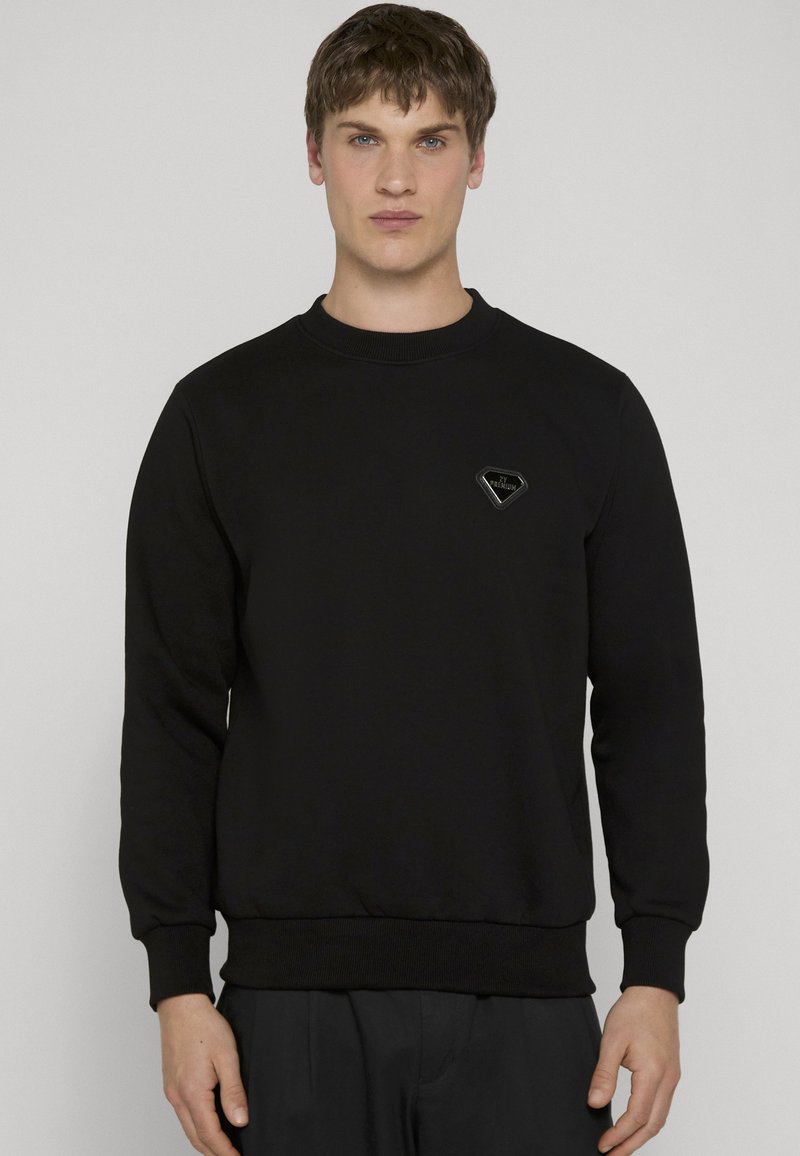 Jeune homme aux cheveux bruns courts portant un sweat-shirt noir à col rond avec un petit logo noir en forme de losange sur la poitrine.