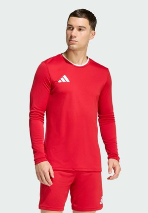 Mann trägt ein rotes langärmeliges Sportshirt und passende Shorts mit weißen Adidas-Logos, steht und schaut zur Seite.