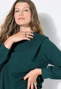 Dunkelgrüner Sweatshirt mit einzigartigem Drapierungsdesign, weichem Stoff und gerippten Bündchen. Das Modell trägt Accessoires: silberne Ohrhänger und Ringe.