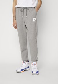 Grå träningsoverallsbyxor med snörning i midjan, sidofickor och en vit lapp med en logotyp. Bärs med vita sneakers med blå och orange detaljer.