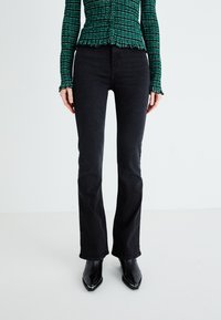 Vaqueros de campana negros con una textura suave y detalles de costura, combinados con botas negras de punta y una blusa verde con estampado.