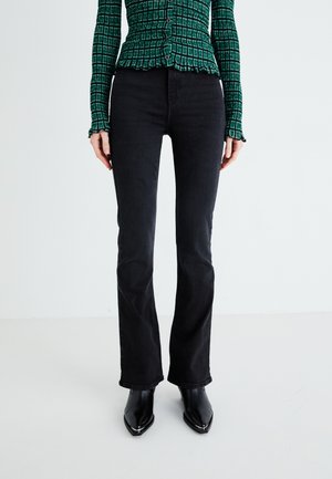 Jeans neri a zampa d'elefante con una texture liscia e dettagli cuciti, abbinati a stivali neri a punta e una maglietta verde con motivo.