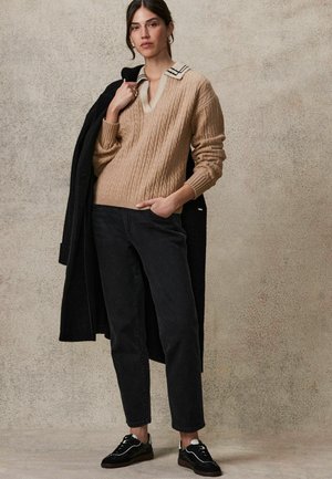 Beige-Strickpullover mit V-Ausschnitt und gestreiftem Kragen, kombiniert mit schwarzen Jeans und schwarz-weißen Sneakers; langer schwarzer Mantel über dem Arm getragen.
