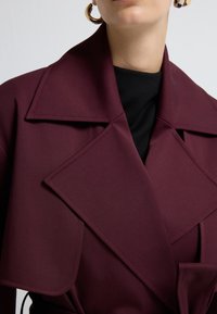 Nærbillede af en person iført en mørk burgunderfrakke med brede klapper over en sort top og guldøreringe mod en ensfarvet baggrund.