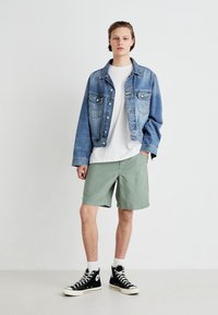 Jachetă din denim de culoare albastru deschis, tricou alb, pantaloni scurți verzi și adidași negri cu vârf înalt. Ținută casual, stratificată, cu culori contrastante.