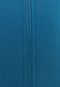 Teal zippered hoodie med en glat, tekstureret stof med en central lynlås, flade sømme og ingen yderligere mønstre eller detaljer.