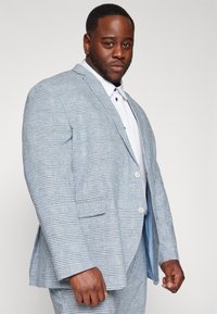 Ανοιχτό μπλε καρό σακάκι με όρθιους γιακάδες, δύο κουμπιά και πλαϊνές τσέπες. Συνδυασμένο με λευκό πουκάμισο με μπλε κουμπιά.