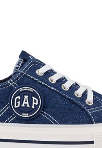 Denim sneaker in dark blue with white stitching. Features round, navy logo patch labeled "GAP" and white laces. Textured fabric and rubber sole.