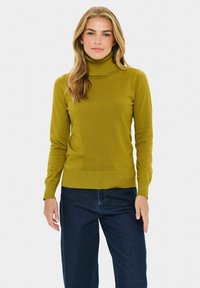 Olivno zeleni pulover s turtleneck ovratom iz rebraste pletenine, z dolgimi rokavi in prilegajočim se krojem, v kombinaciji s temno modrimi širokimi kavbojkami.