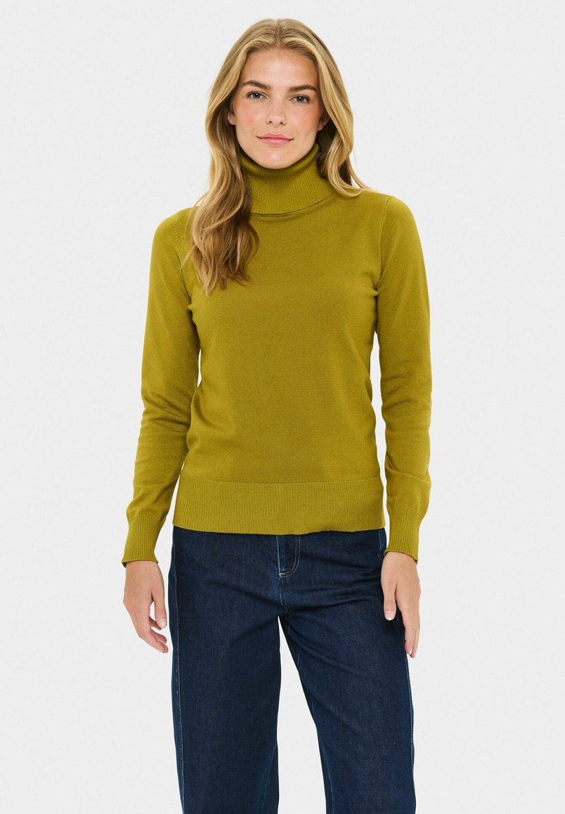 Olivno zeleni pulover s turtleneck ovratom iz rebraste pletenine, z dolgimi rokavi in prilegajočim se krojem, v kombinaciji s temno modrimi širokimi kavbojkami.