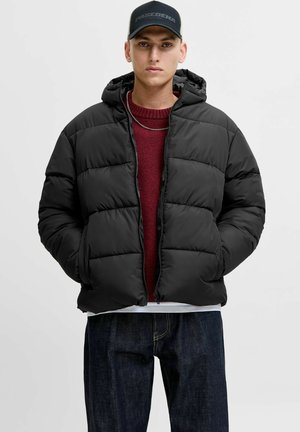 Chaqueta acolchada negra con capucha, con un diseño acolchado. Llevada sobre un suéter burdeos y combinada con jeans oscuros.