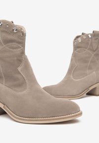 Bottes en daim beiges à cheville avec des coutures décoratives, des découpes sur les côtés et des accessoires en métal argenté. Talon bas empilé et semelle en caoutchouc plate.