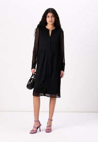 Μαύρο φόρεμα με κουμπωτή μπροστινή όψη, μακρύς διαφανής μανίκι και συγκεντρωμένη μέση. Συμπληρωμένο με μοβ ψηλές γόβες με straps και μια μικρή μαύρη τσάντα.