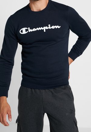 Muž v tmavě modré mikině značky Champion a tmavě šedých teplácích Puma stojí s jednou rukou na bok proti bílému pozadí.