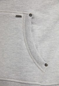 Grau Sweatshirtstoff mit einer weichen Textur, ausgestattet mit einer Tasche mit ribbedem Besatz und metallischen Akzenten. Die Nähte sind entlang der Kanten sichtbar.