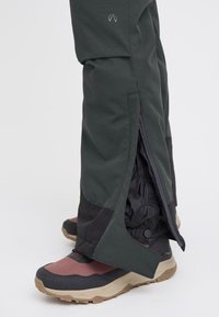 Mørkegrønne vanntette bukser med svarte detaljer og sidelommer, kombinert med flerfargede turstøvler med en teksturert såle.