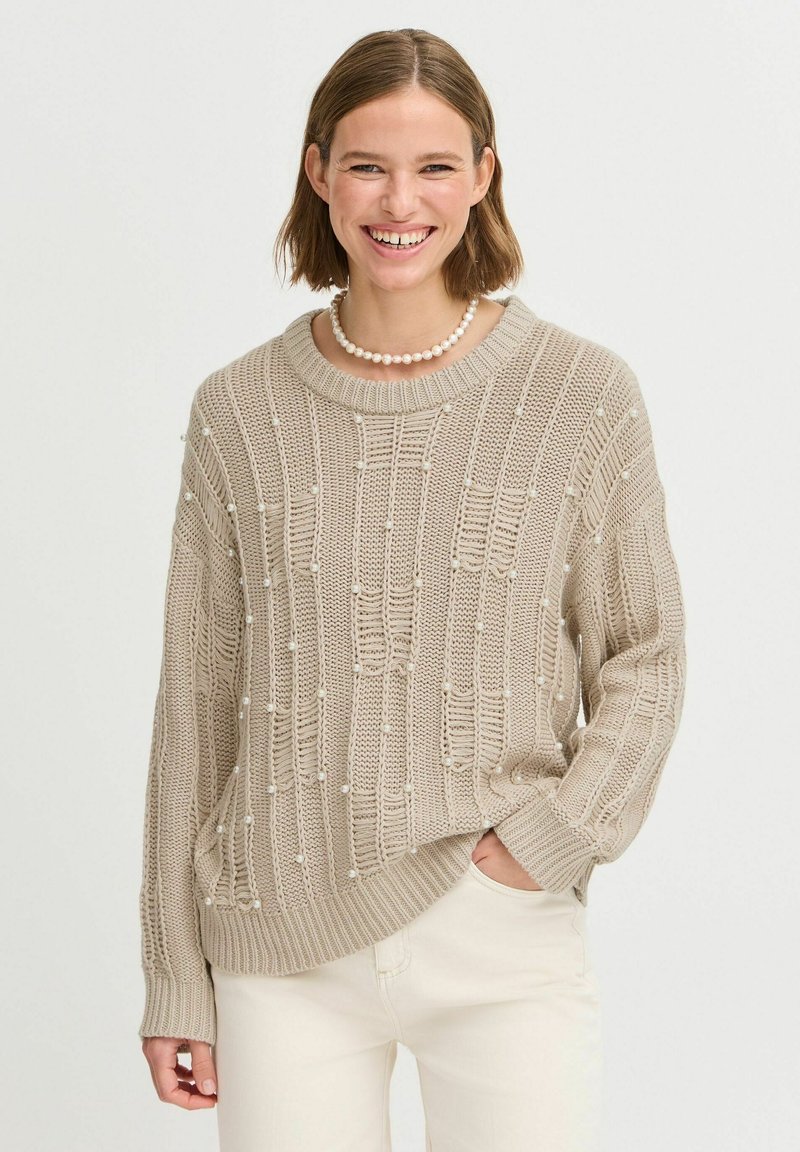 Pull en maille beige avec un motif texturé et des détails en relief, agrémenté de petites perles blanches. Coupe ample avec col et poignets côtelés.
