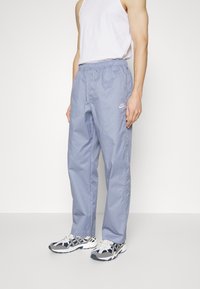 Pantaloni da pista Nike azzurro chiaro con vita elastica e tasche laterali, con un piccolo logo bianco sulla coscia sinistra. Indossati con scarpe da ginnastica.