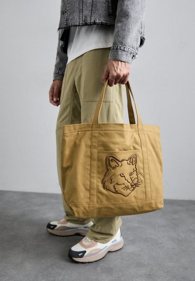 Geantă tote din pânză bej, cu o broderie în formă de cap de vulpe. Dispune de mânere rezistente și o formă rectangulară, asortată cu pantaloni cargo de culoare verde măslin.