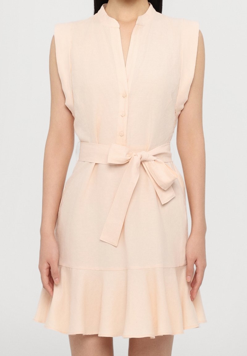 Robe sans manches d'un pêche clair avec un col rond, des boutons sur le devant, une ceinture nouée et un ourlet froncé. Fabriquée en tissu doux et texturé.