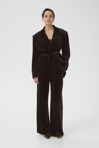 Černý sametový blejzr s páskem v pase, velkými klopami a přední kapsou, kombinovaný s širokými kalhotami a špičatými zelenými podpatky.