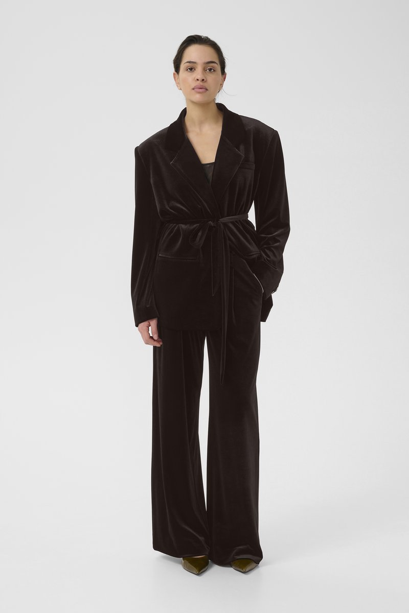 Schwarzer Samtblazer mit tailliertem Gürtel, großen Revers und einer Fronttasche, kombiniert mit weit geschnittenen Hosen und spitzen grünen Pumps.