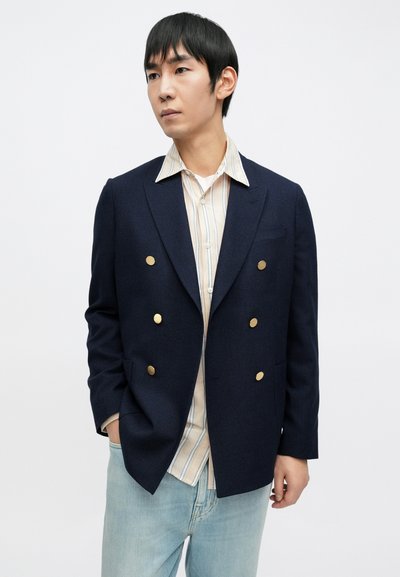 Granatowa marynarka dwurzędowa ze złotymi guzikami, z teksturowanej tkaniny, w połączeniu z koszulą w paski i jasnoniebieskimi jeansami.